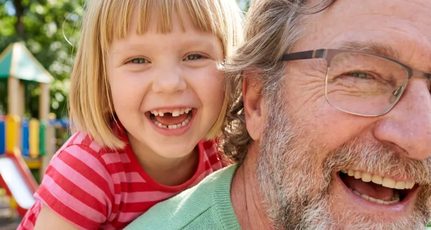 Senior aktivoptik - Nahaufnahme kleines Mädchen mit Zahnlücke und älterer Mann mit Brille, welcher nur zur Hälfte abgebildet ist, sind lachend auf einem Spielplatz