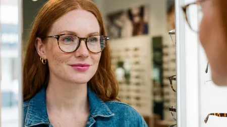 Brillenformen aktivoptik - Frau mit Brille und dünnen rund geformten Augenbrauen schaut lächelnd in einen Spiegel in einem Optikgeschäft