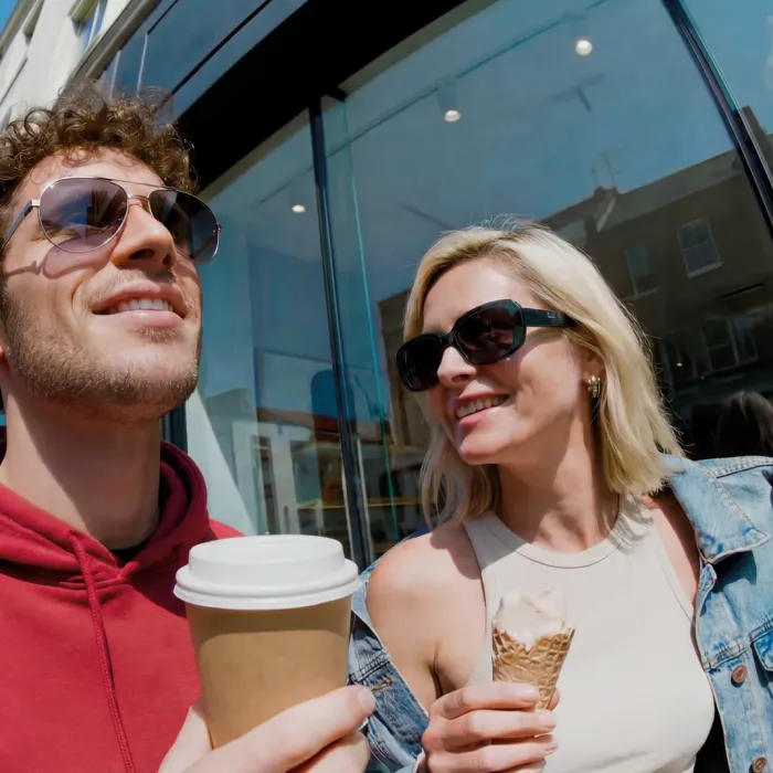 Polarisierende Gläser aktivoptik - Frau und Mann mit Sonnenbrille und Kaffe laufen an einem Schuaufenster vorbei während die Frau den Mann anschaut 